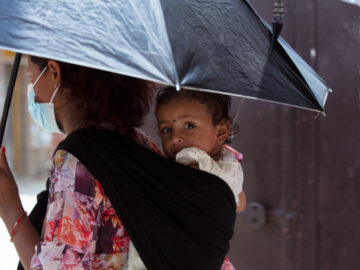 A photo of a woman holding an umbrella and carrying a baby in a sling on her back. The photo was taken in Nepal, which is one of many countries at risk of typhoid related to climate change and extreme weather.