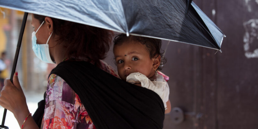 A photo of a woman holding an umbrella and carrying a baby in a sling on her back. The photo was taken in Nepal, which is one of many countries at risk of typhoid related to climate change and extreme weather.